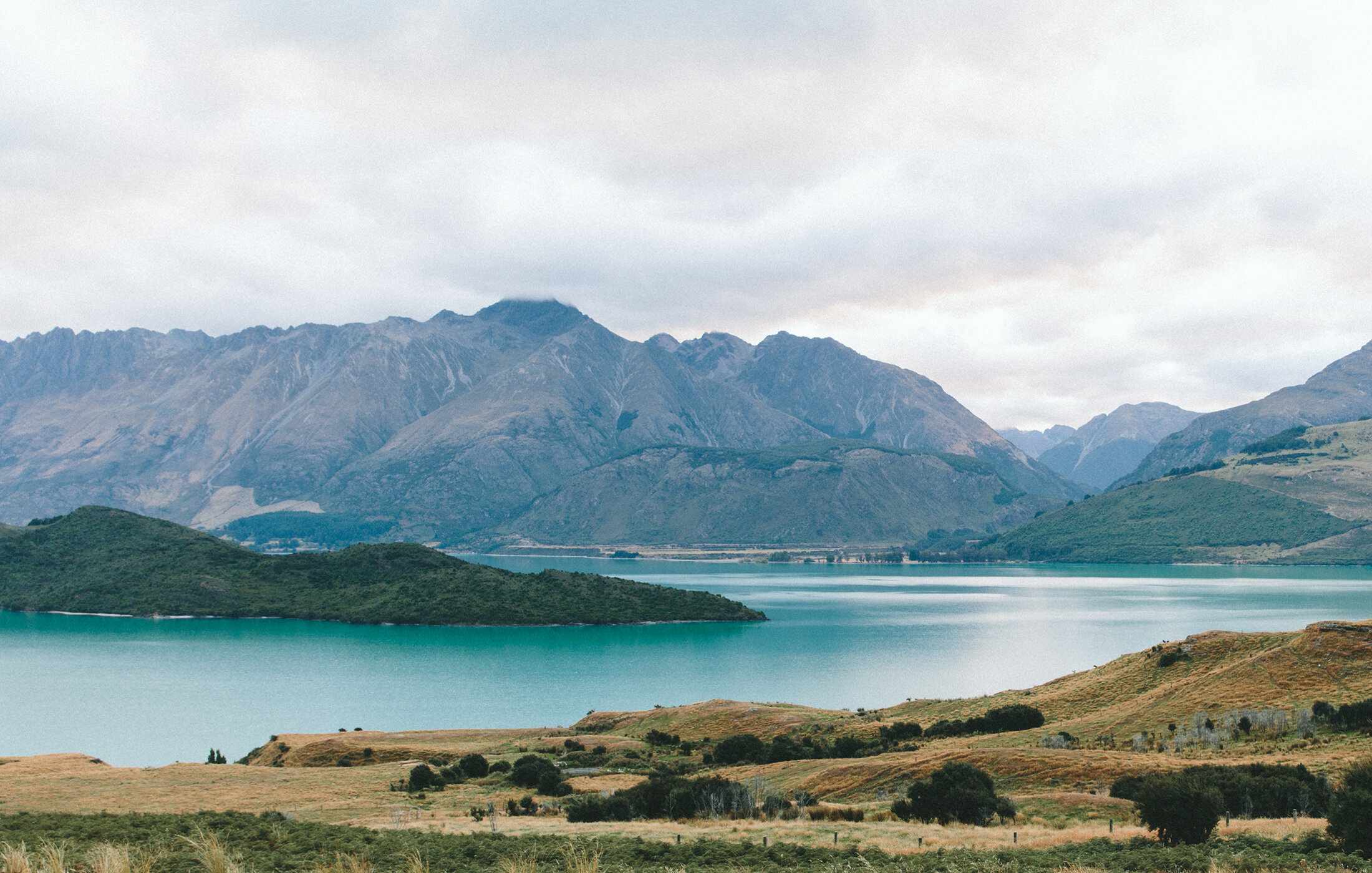 A scenic view of a calm lake bordered by lush green hills and distant rugged mountains under a cloudy sky.