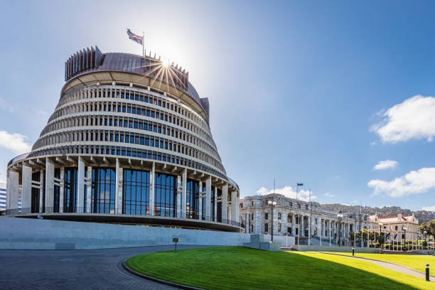 The image shows the Beehive building in Wellington, New Zealand, with the sun shining above it and a clear blue sky.