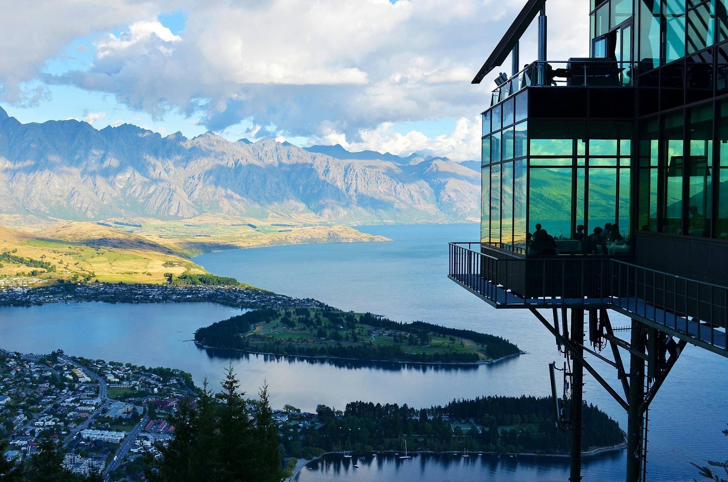 A modern glass building, compliant with the Resource Management Act, overlooks a scenic landscape with a lake, small islands, and mountains under a partly cloudy sky.